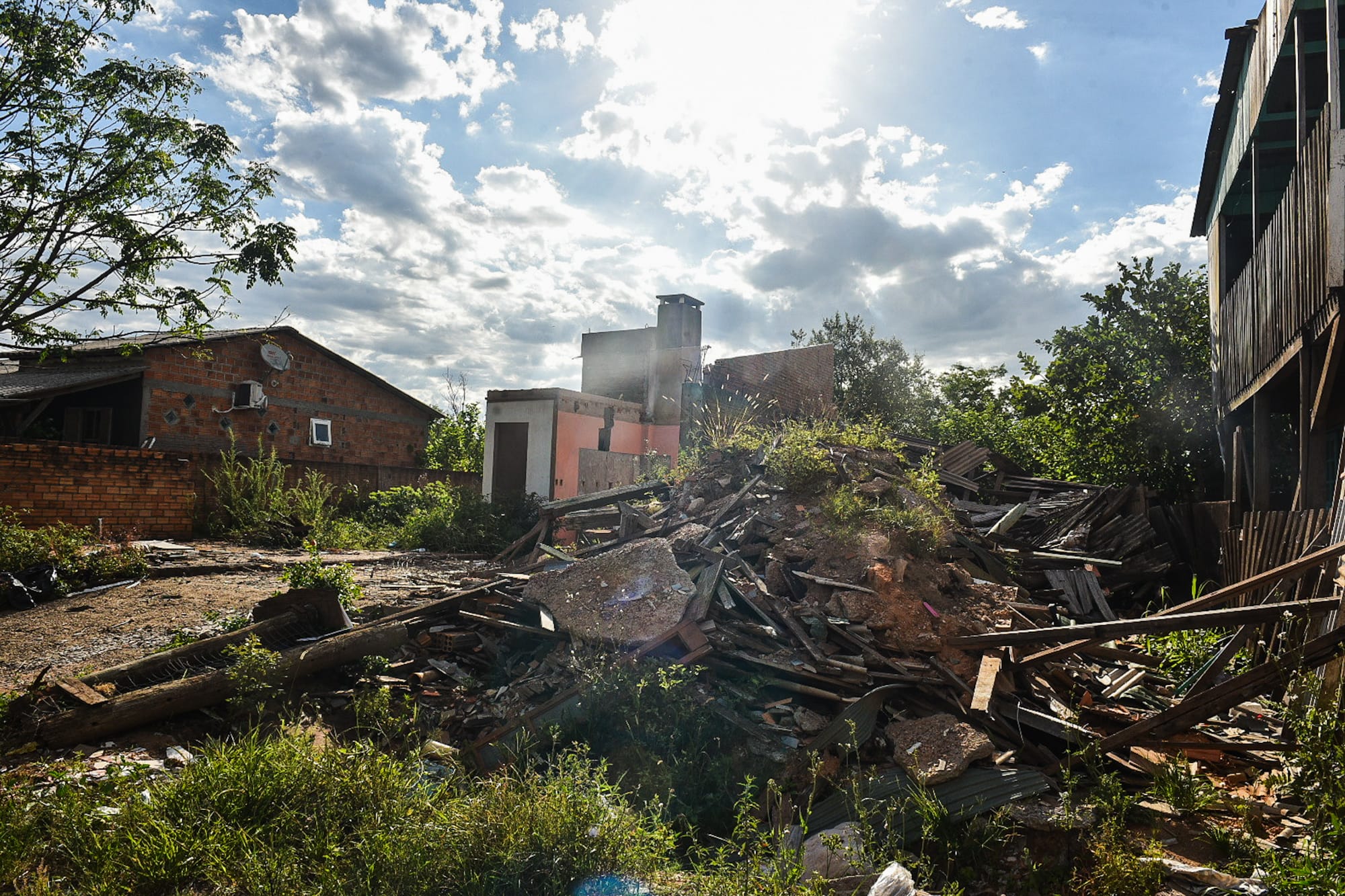 Pilha de entulho e restos de construção em terreno entre casas simples, com sol forte e céu parcialmente nublado ao fundo.