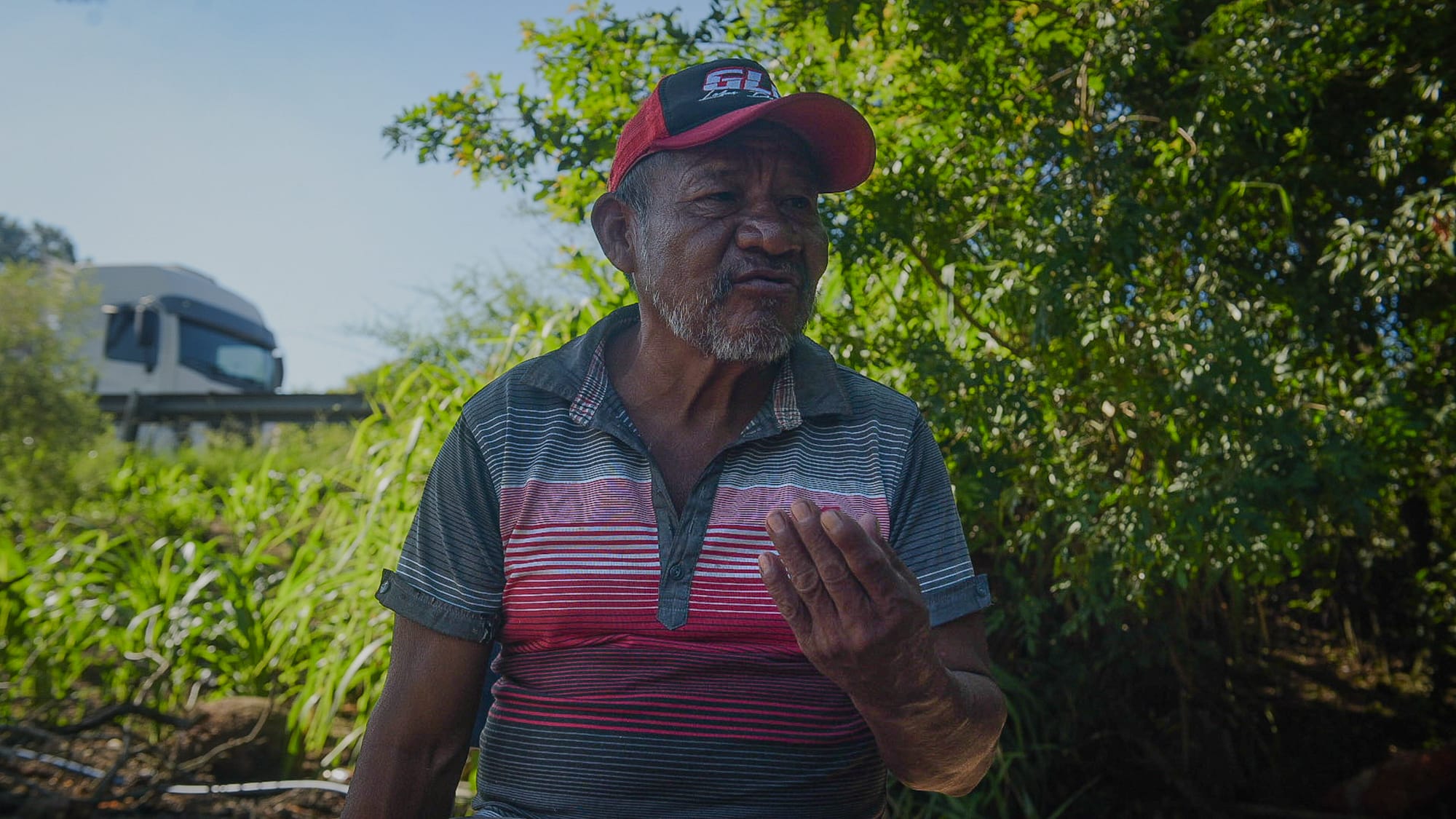 Homem idoso com boné vermelho e camisa listrada fala gesticulando com a mão, em área verde próxima a uma estrada.
