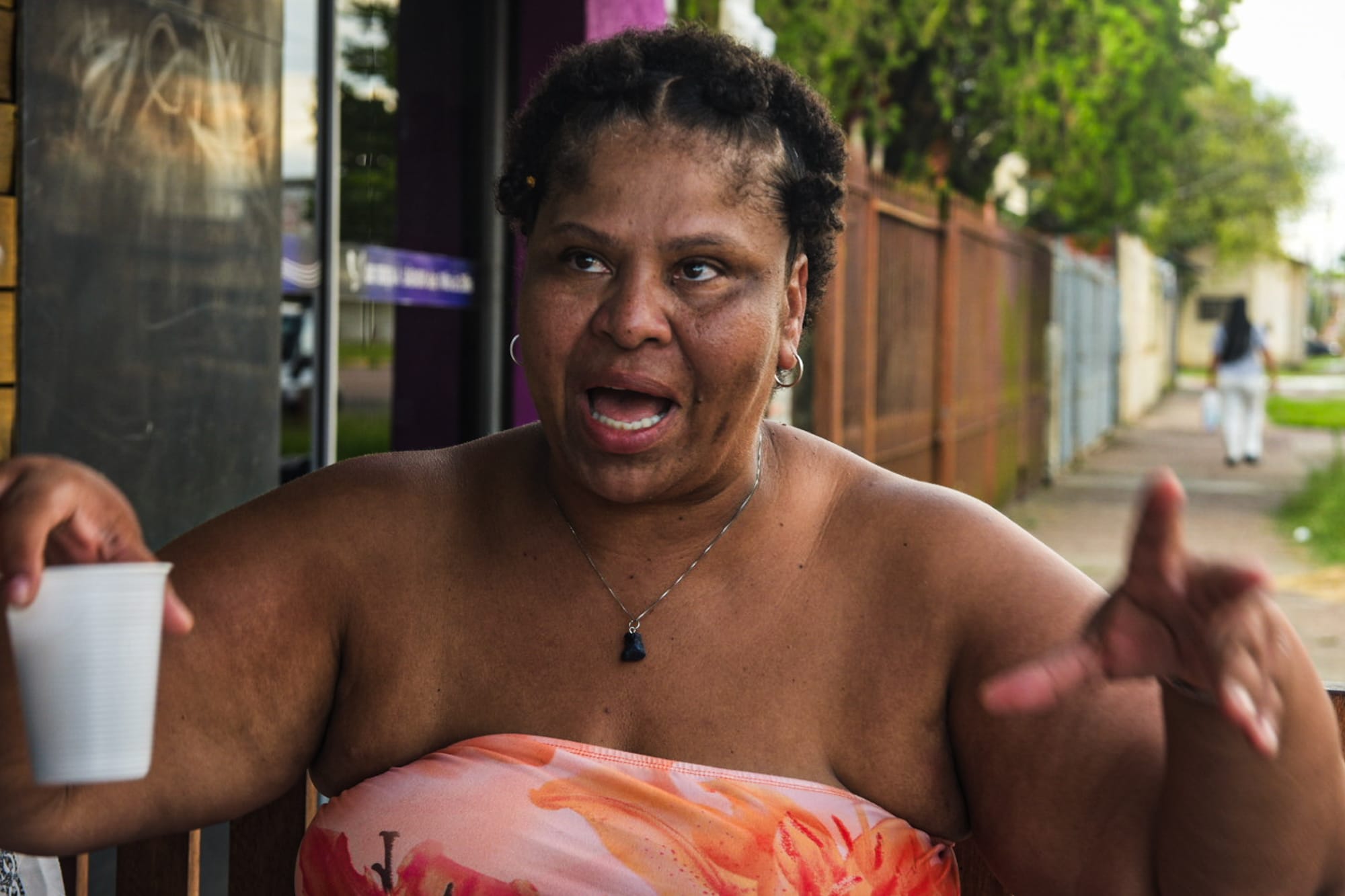 Mulher sentada na calçada segura um copo descartável e gesticula enquanto fala, com rua e casas ao fundo.